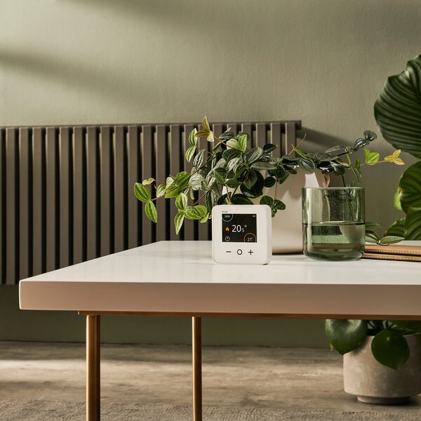 A white Wiser smart room thermostat displaying 20.5°C sitting on a table in front of a radiator