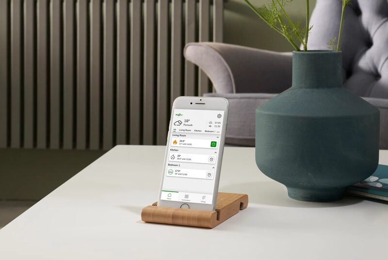 A smartphone on a wooden stand displaying the Wiser smart heating control app, with room temperatures, next to a vase on a table with a radiator behind