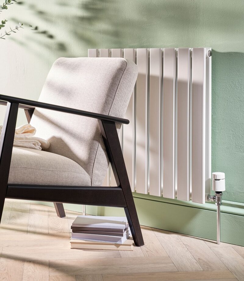 A white vertical-panel radiator with a smart thermostatic valve fitted, in a living room beside an armchair