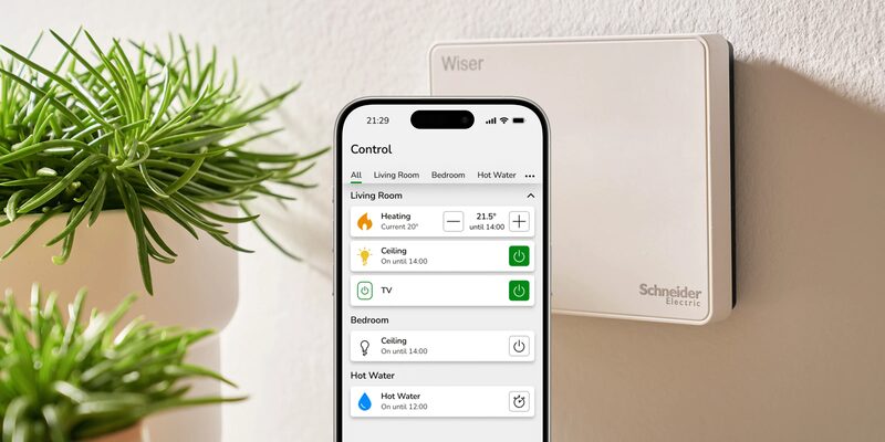 A Wiser smart heating controller mounted on a wall next to a smartphone showing the Wiser app's heating control screen