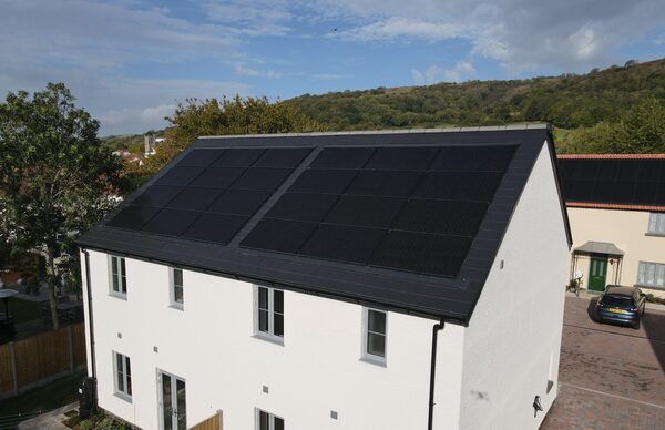 A white two-storey house with black solar panels covering the roof, in a UK village setting