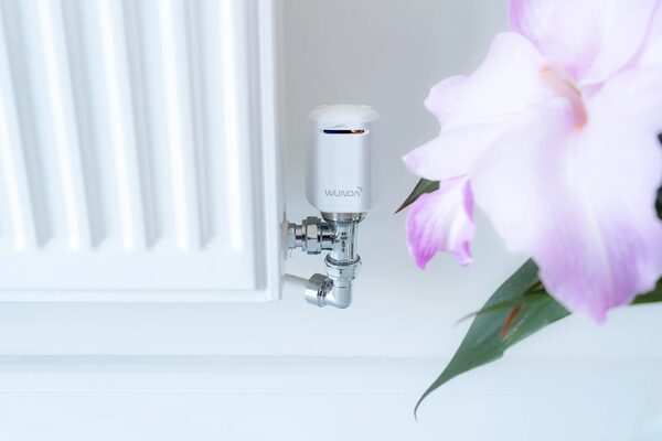 A white Wunda smart thermostatic radiator valve head fitted to a radiator valve beside a pink flower