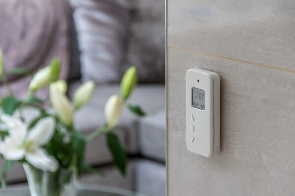 A white WundaSmart room thermostat mounted on a tiled wall showing 21.0°C, with a blurred living room and flowers in the background