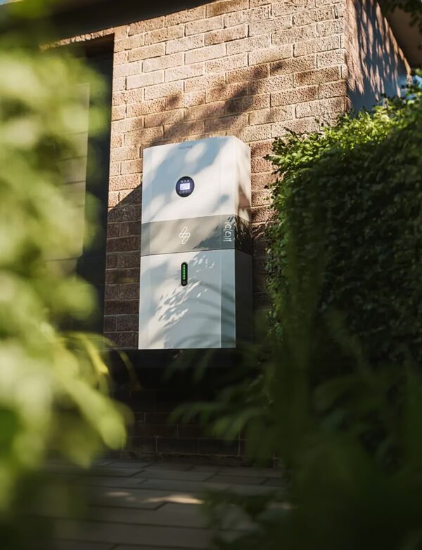 A myenergi libbi home battery storage unit mounted on the brick exterior wall of a house beside a hedge