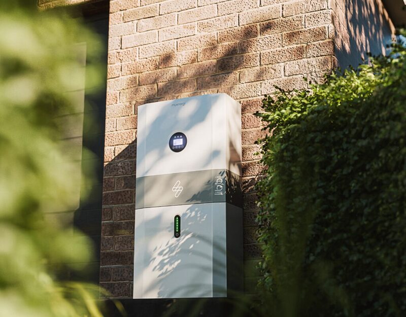 A myenergi libbi home battery storage unit mounted on an exterior brick wall of a house beside a hedge
