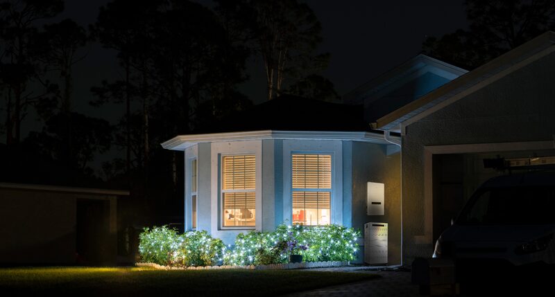 A house at night with a wall-mounted home battery unit glowing beside the garage