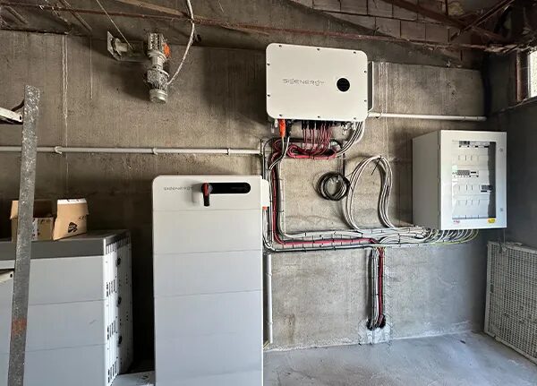 A Sigenergy home battery and inverter installation with wiring and a consumer unit mounted on a bare concrete wall in an unfinished utility space