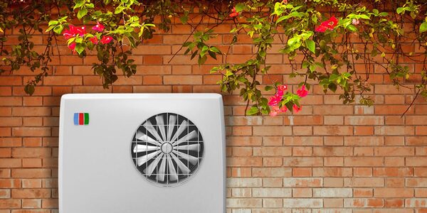 A white air source heat pump outdoor unit with a fan grille mounted against a red brick wall with flowering plants above