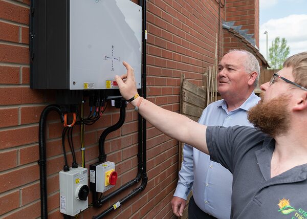 An installer pointing at a wall-mounted battery storage unit outside a brick house while a homeowner looks on