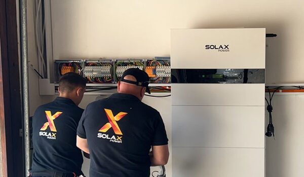 Two installers in SolaX Power branded shirts working beside a wall-mounted SolaX home battery next to electrical distribution boards