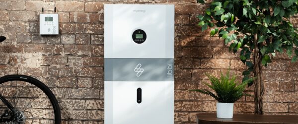 A white myenergi libbi home battery storage unit mounted against a brick wall with a small controller beside it and a plant nearby