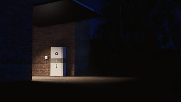 A myenergi libbi home battery storage unit mounted on a brick wall outside a house at night