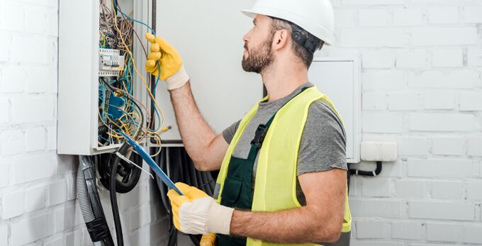 An electrician in a hard hat and hi-vis vest inspecting the wiring in an open consumer unit while holding a clipboard