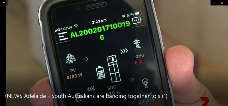 A hand holding a smartphone displaying an Alpha ESS battery monitoring app showing solar PV, battery state of charge and grid power flows