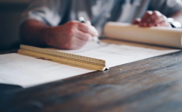 A person signing or drawing on paper plans at a desk with a wooden ruler and rolled-up drawings