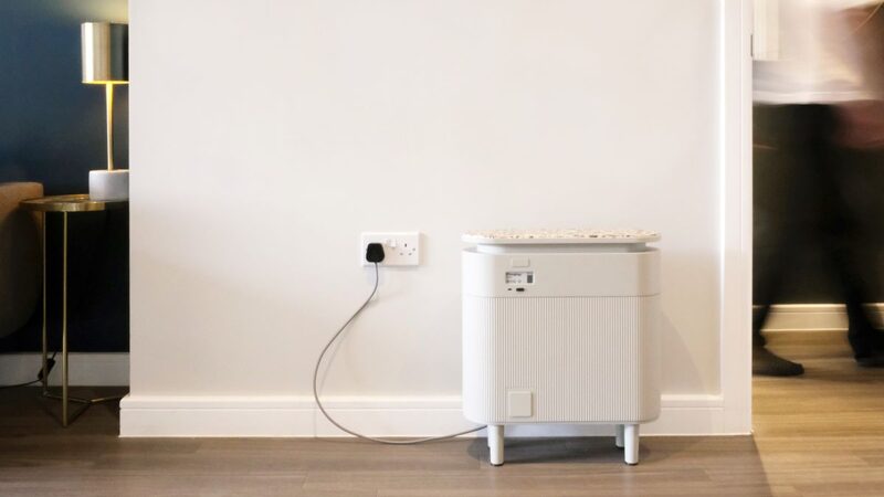 A white plug-in home battery unit on legs, plugged into a wall socket in a home hallway