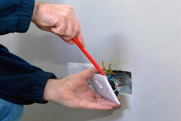 An electrician fitting a white UK double socket onto exposed wiring in a wall using a red screwdriver
