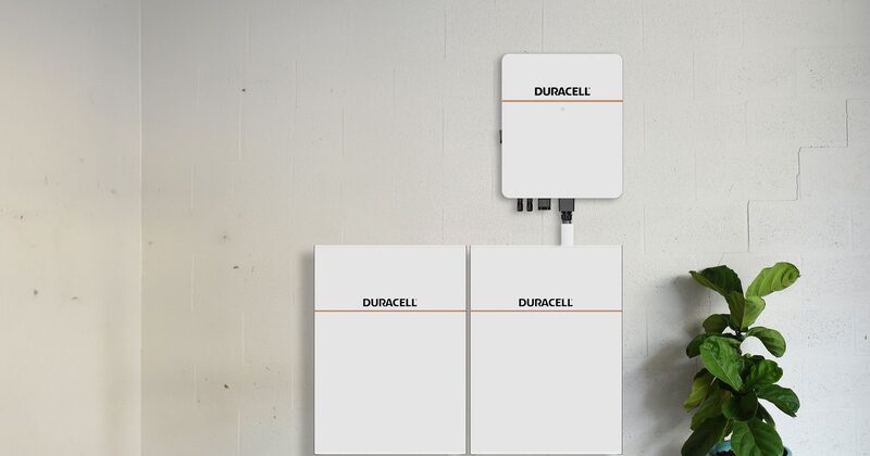 Two Duracell home battery storage units on the floor with a wall-mounted Duracell inverter above them next to a potted plant