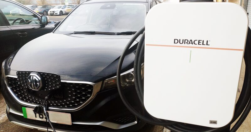 A Duracell-branded EV wall charger with cable plugged into a black MG electric car