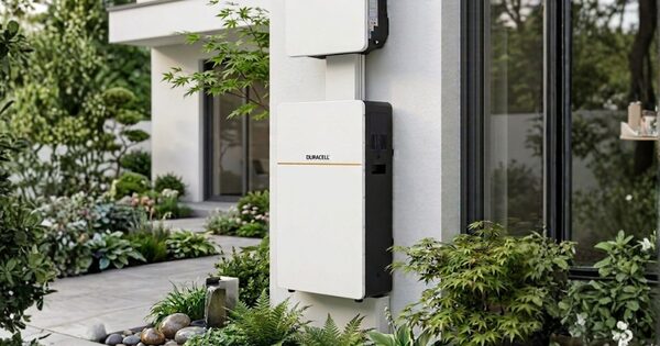 A white Duracell Dura16 home battery mounted on an exterior house wall beside a garden