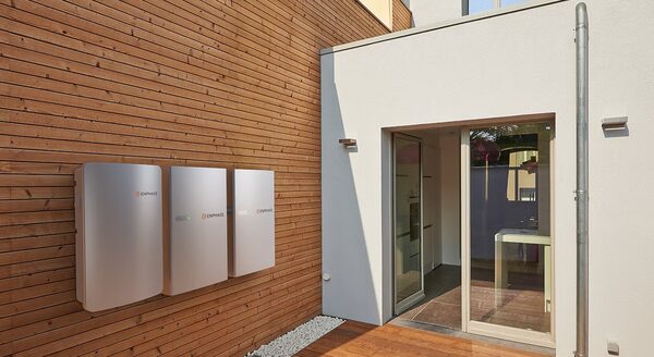 Three Enphase home battery units mounted on a timber-clad exterior wall beside a modern house entrance