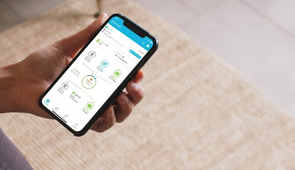 A hand holding a smartphone displaying the Enphase app home energy monitoring dashboard