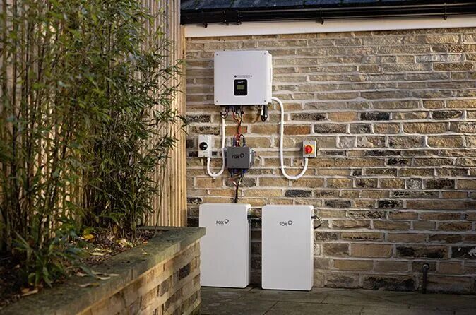 A Fox solar battery storage system with two white battery units and an inverter mounted on an outdoor brick wall of a house