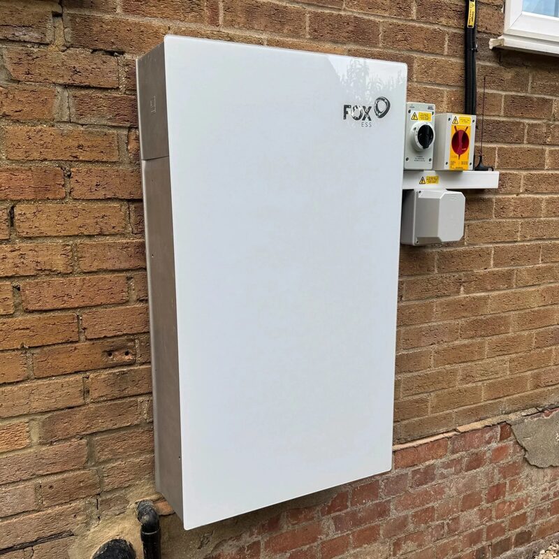 A white Fox ESS battery storage unit mounted on an exterior brick wall next to electrical isolation switches