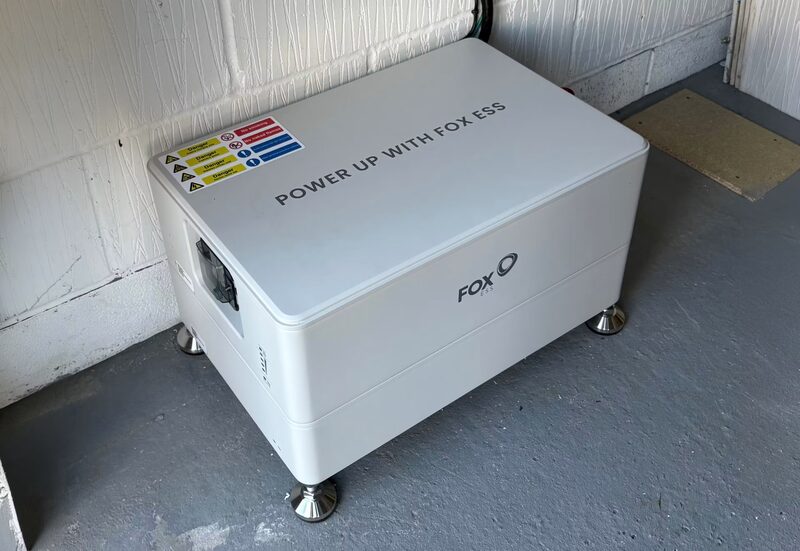 A white Fox ESS home battery installed on the floor of a garage against a painted brick wall
