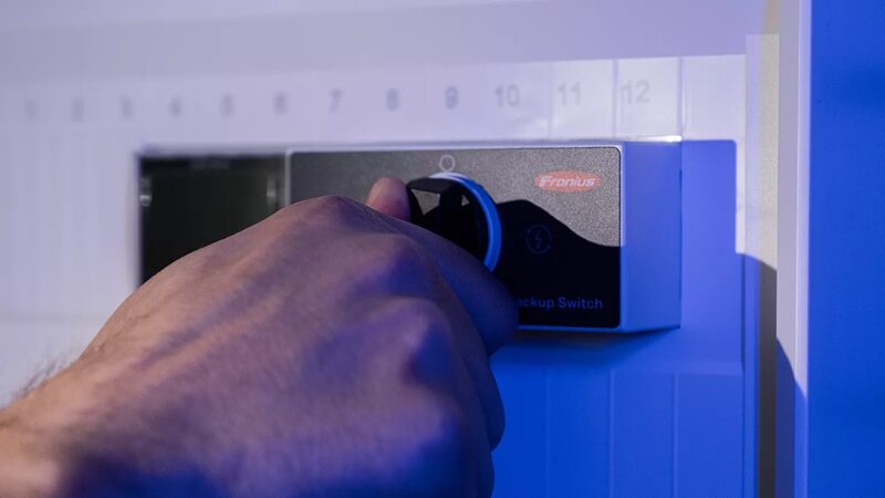 A hand turning the dial on a Fronius backup switch mounted on a wall distribution board