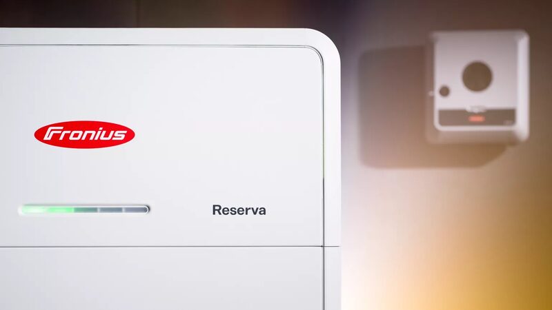 A close-up of a white Fronius Reserva home battery storage unit with a green charge indicator, mounted next to a wall box
