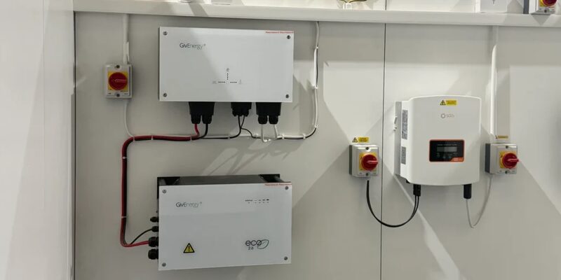 A GivEnergy battery inverter and battery unit wall-mounted next to a Solis solar inverter with red emergency switches, on a white wall