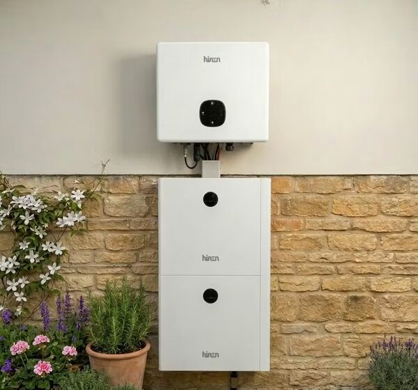 A white Hinen home battery and inverter unit mounted on an outside stone wall of a house, with garden plants beside it