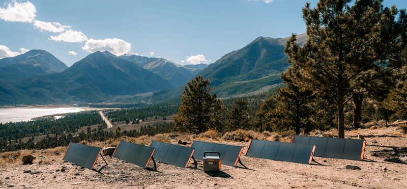 A Jackery portable power station connected to several folding solar panels set up on a hillside in a mountain landscape