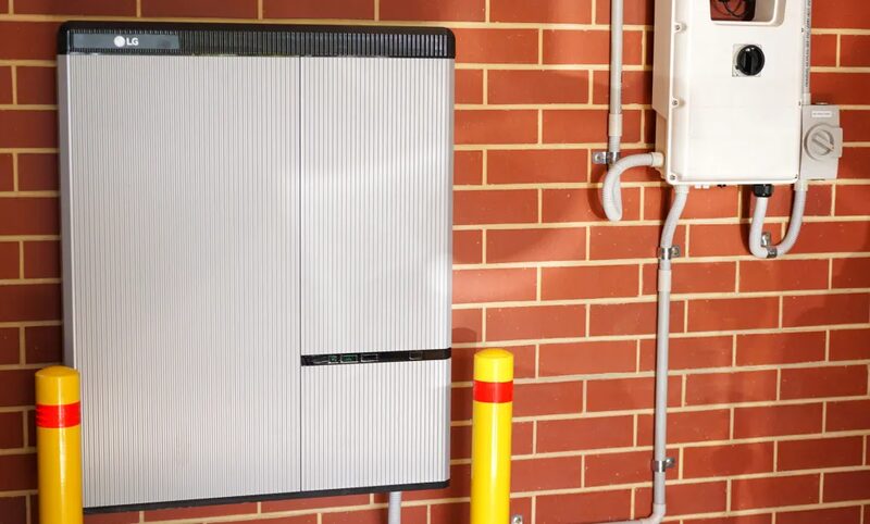 An LG domestic battery storage unit mounted on a red brick wall next to an inverter and yellow safety bollards