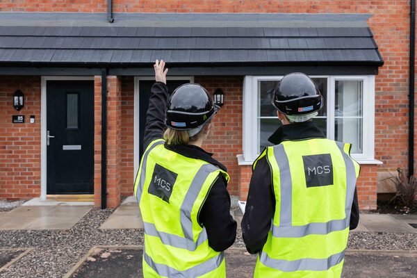 Two installers in hi-vis MCS vests and hard hats pointing at the roof of a brick house