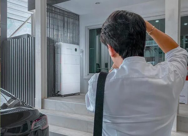 A man photographing a white Sigenergy home battery unit installed on a porch next to a parked car