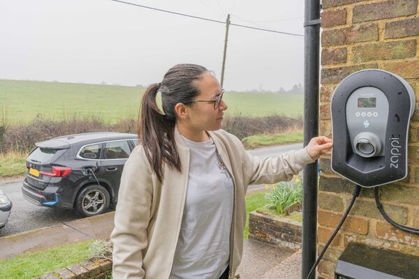 A woman using a myenergi Zappi EV charger mounted on a brick house wall with an electric car parked outside