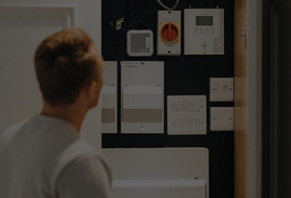 A man looking at a wall of electrical consumer units and a myenergi libbi battery storage controller and harvi unit installed in a home