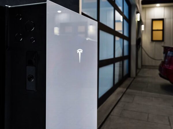 A white Tesla Powerwall 3 home battery installed on a wall outside a house next to a garage with an EV charging cable