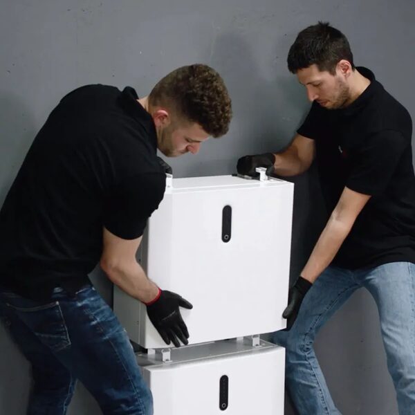 Two installers in black gloves mounting a white SolarEdge home battery unit onto a stacked battery rack against a grey wall