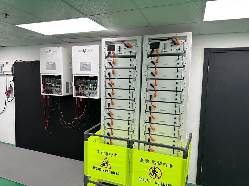 Two Sunsynk inverters mounted on a wall next to two tall stacks of battery modules in an indoor room, with yellow work-in-progress barriers in front
