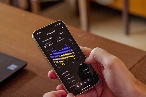 A hand holding a smartphone displaying the Tesla Powerwall energy monitoring app with charge and energy flow charts