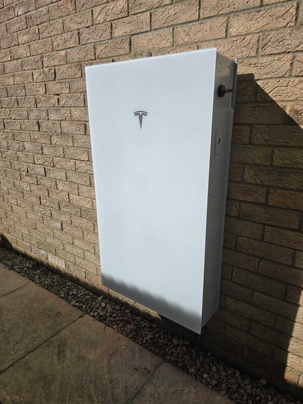 A white Tesla Powerwall home battery mounted on an exterior brick wall of a house