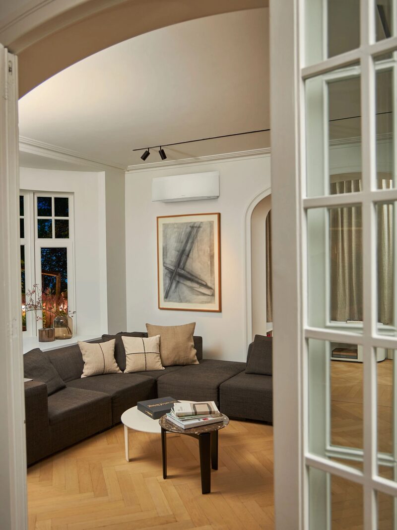 A wall-mounted air-to-air heat pump unit installed above a grey corner sofa in a bright living room