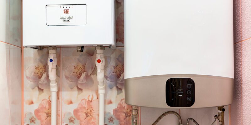 Two white electric boilers (a wall-mounted combi unit and a cylinder-style unit) installed side by side on a floral-tiled wall with pipework