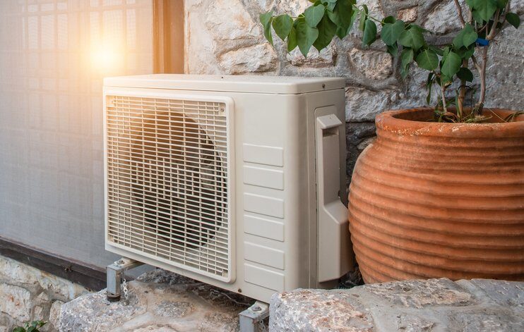 An air source heat pump outdoor unit installed against a stone wall beside a large terracotta plant pot