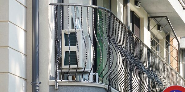 A white air source heat pump unit installed on a residential balcony behind a decorative black metal railing