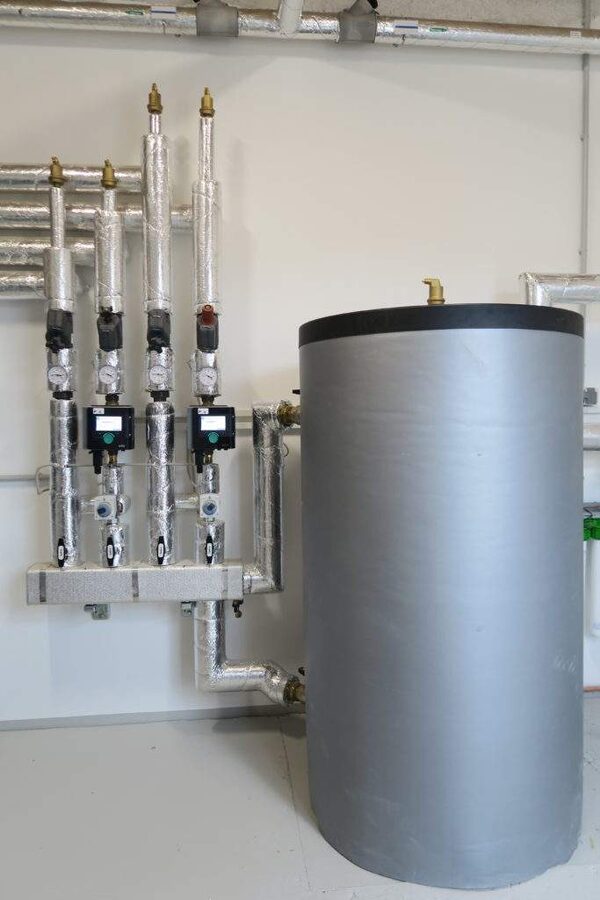A large grey insulated hot water cylinder connected to insulated pipework, valves and pump manifolds in a plant room