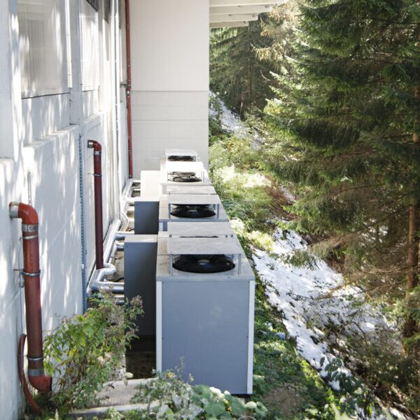 A row of grey heat pump outdoor units with fans installed along the exterior wall of a building beside trees